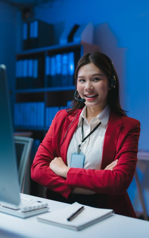 Confident asian female call center operator wearing headset smiling with arms crossed while working late night shift at office desk
