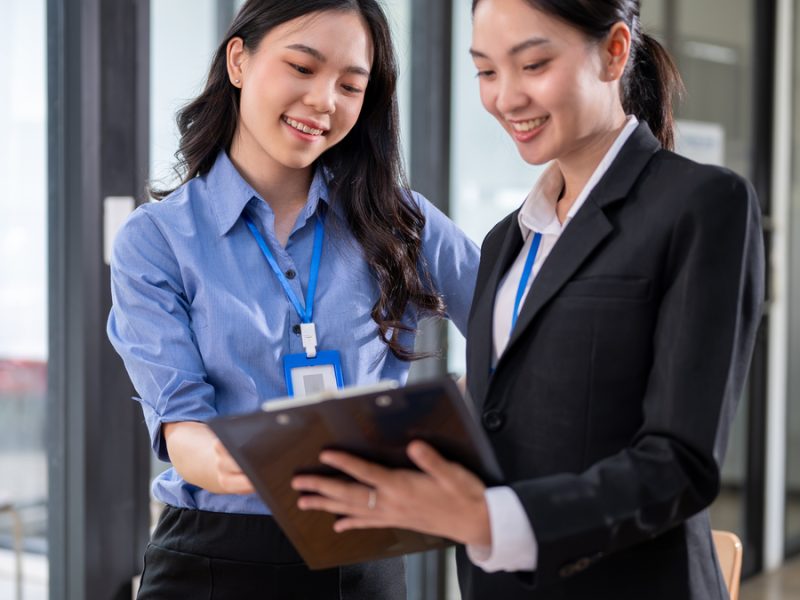 Two happy and beautiful millennial Asian businesswomen are having a discussion and examining business documents on a clipboard while standing in the office together. colleagues, coworkers, teamwork