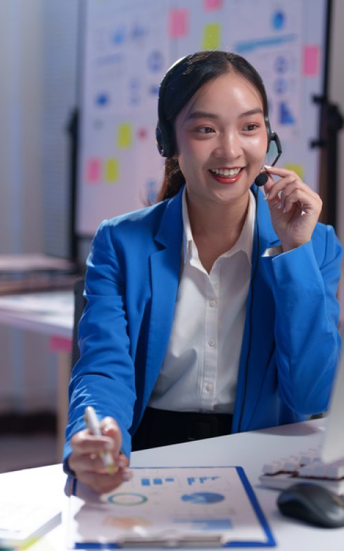 Young professional woman in blue jacket smiling while working late at night in office using headset and computer, taking notes on clipboard with charts and diagrams